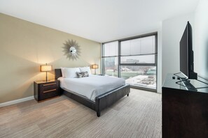 A modern hotel room with a large bed, two bedside tables with lamps, a TV, and a view of the city.