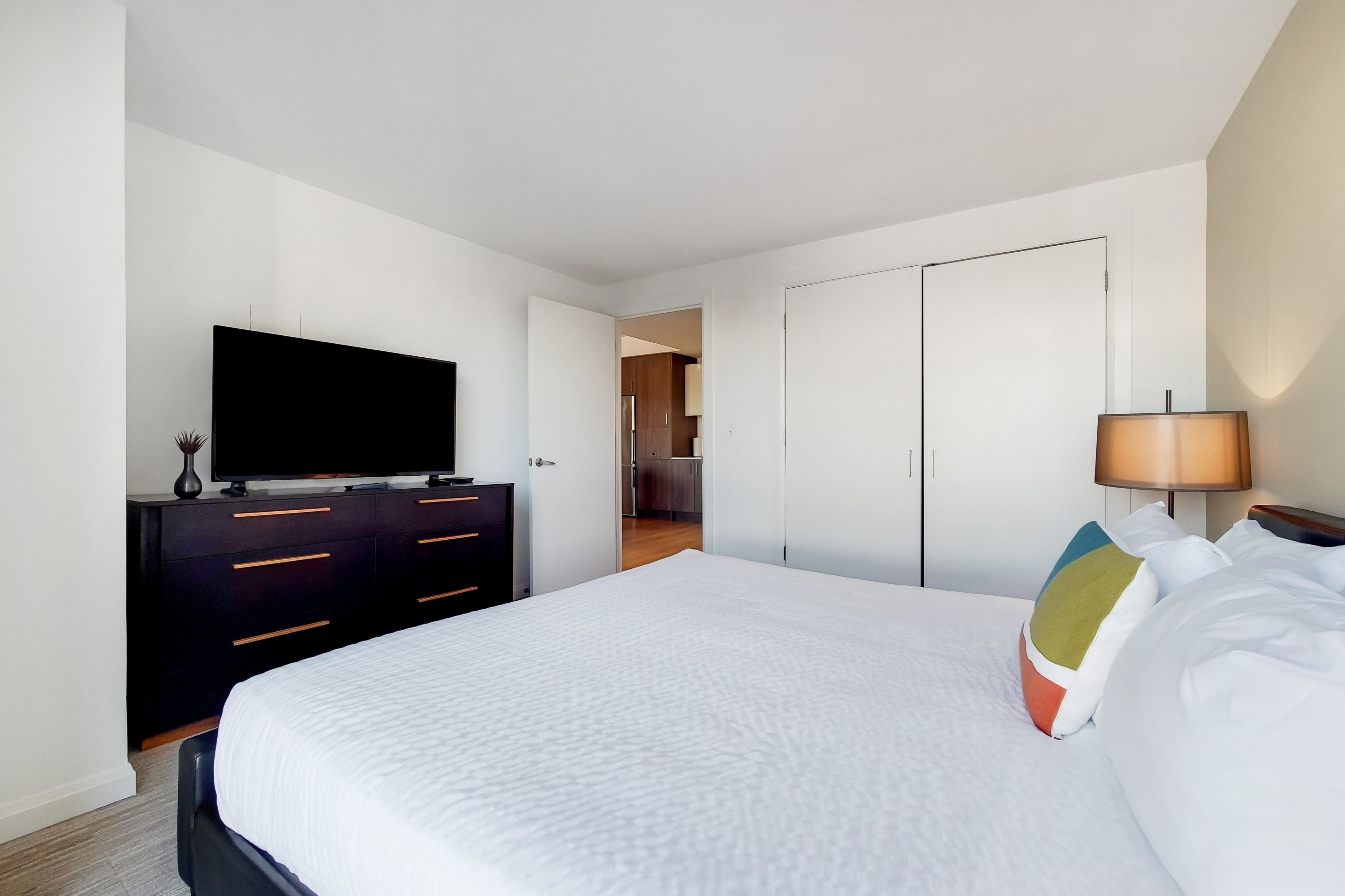 A hotel room with a large bed, a TV on a dresser, a lamp, and a door leading to a bathroom.