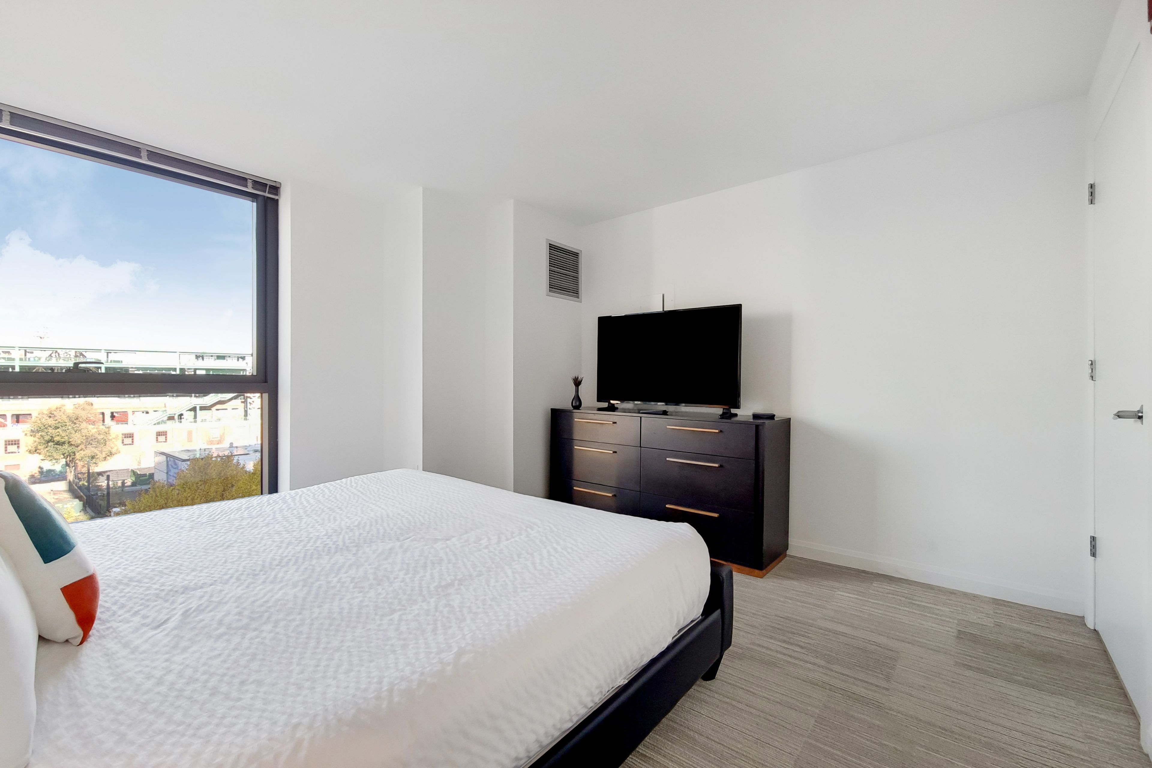 A bedroom with a large window, a bed, a TV on a black dresser, and a city view.