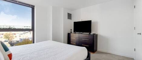 A bedroom with a large window, a bed, a TV on a black dresser, and a city view.