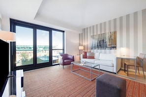 A modern living room with a large window, a white sofa, a red armchair, a glass coffee table, and a striped wall. - Serviced 1 Bedroom Across From Fenway Park (Boston)
