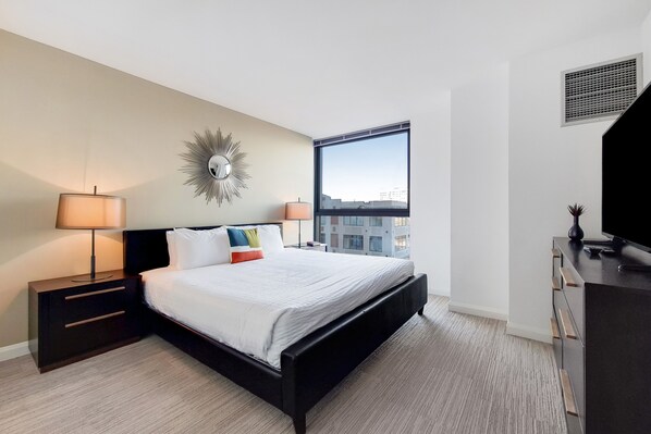 Habitación de hotel moderna con una cama grande, dos mesitas de noche con lámparas, un aparador y un amplio ventanal con vistas a la ciudad.