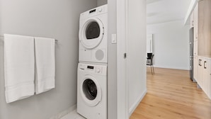 A modern laundry room with stacked washers and dryers, a wooden floor, and a view into the adjacent living area. - Spacious One Bedroom, Steps from Fenway Park (Boston)