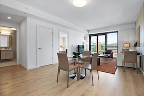 A modern living room with a glass dining table, comfortable seating, and a large window offering a city view.