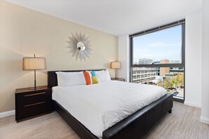 A modern hotel room with a large bed, two bedside tables, a mirror, and a view of the city through a large window.
