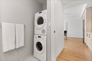 A modern laundry room with stacked washers and dryers, a wooden floor, and a view into the adjacent living area.