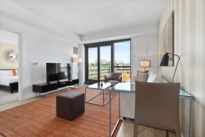 A modern hotel room with a large window, a flat-screen TV, a glass coffee table, a sofa, and a desk with a lamp. - Spacious One Bedroom, Steps from Fenway Park (Boston)