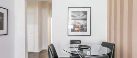 A modern dining area with a glass table and black chairs, framed artwork on the wall, and a door leading to another room.