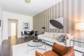 A modern living room with a white sofa, leather armchair, and glass coffee table. There is a dining area with a table and chairs, and a large abstract painting on the wall.