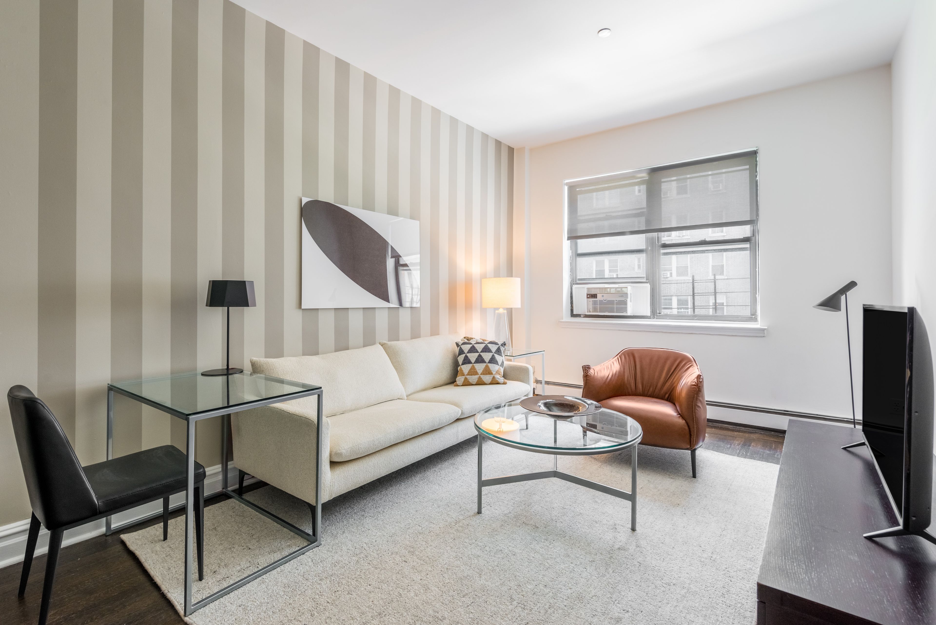 A modern living room with a glass coffee table, a sofa, a leather armchair, a TV, and a large window.