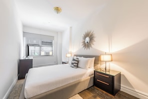 A modern bedroom with a large bed, bedside lamps, a decorative mirror, and a view of the outside through a window.