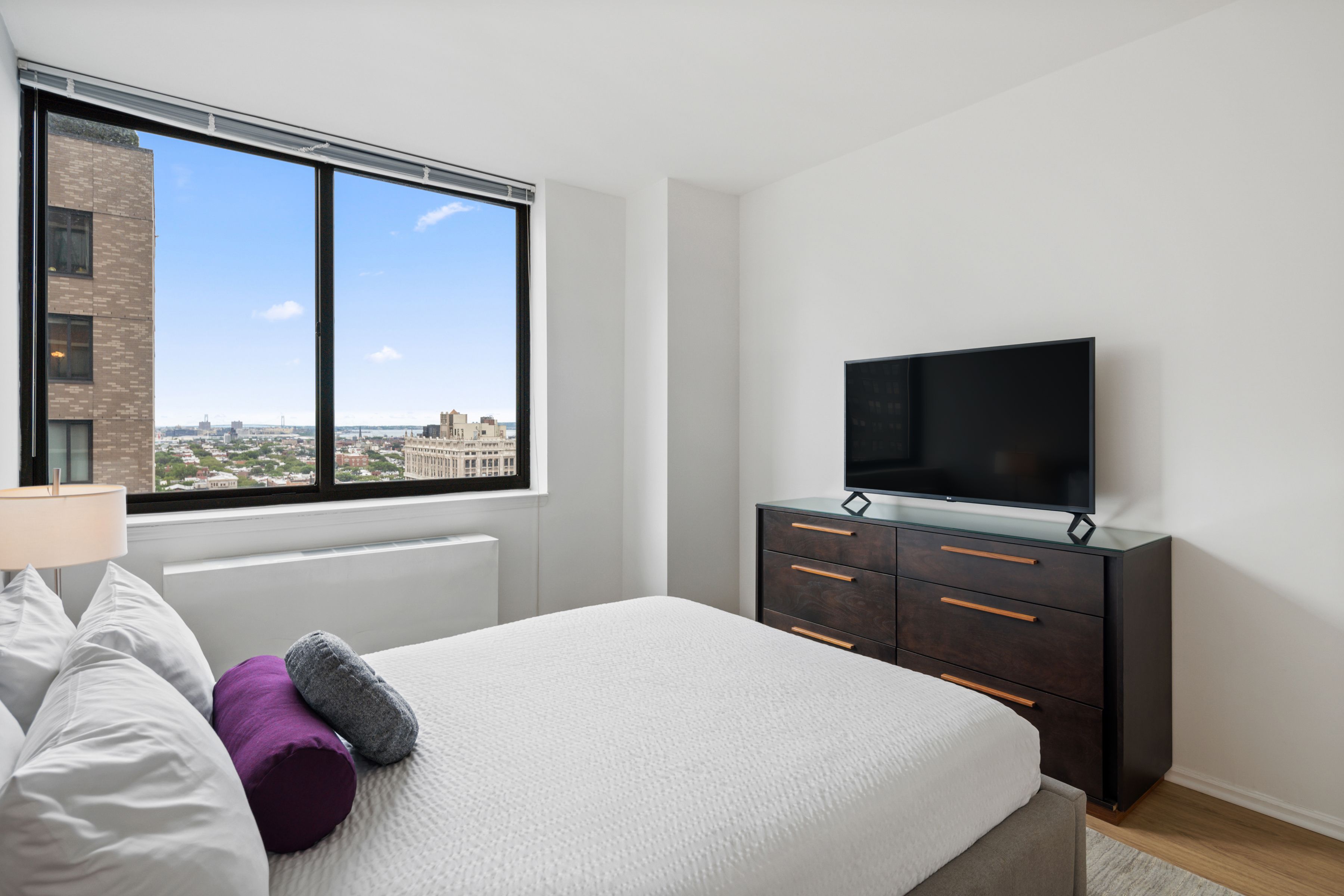 A bedroom with a bed, a dresser, a television, and a window with a city view.