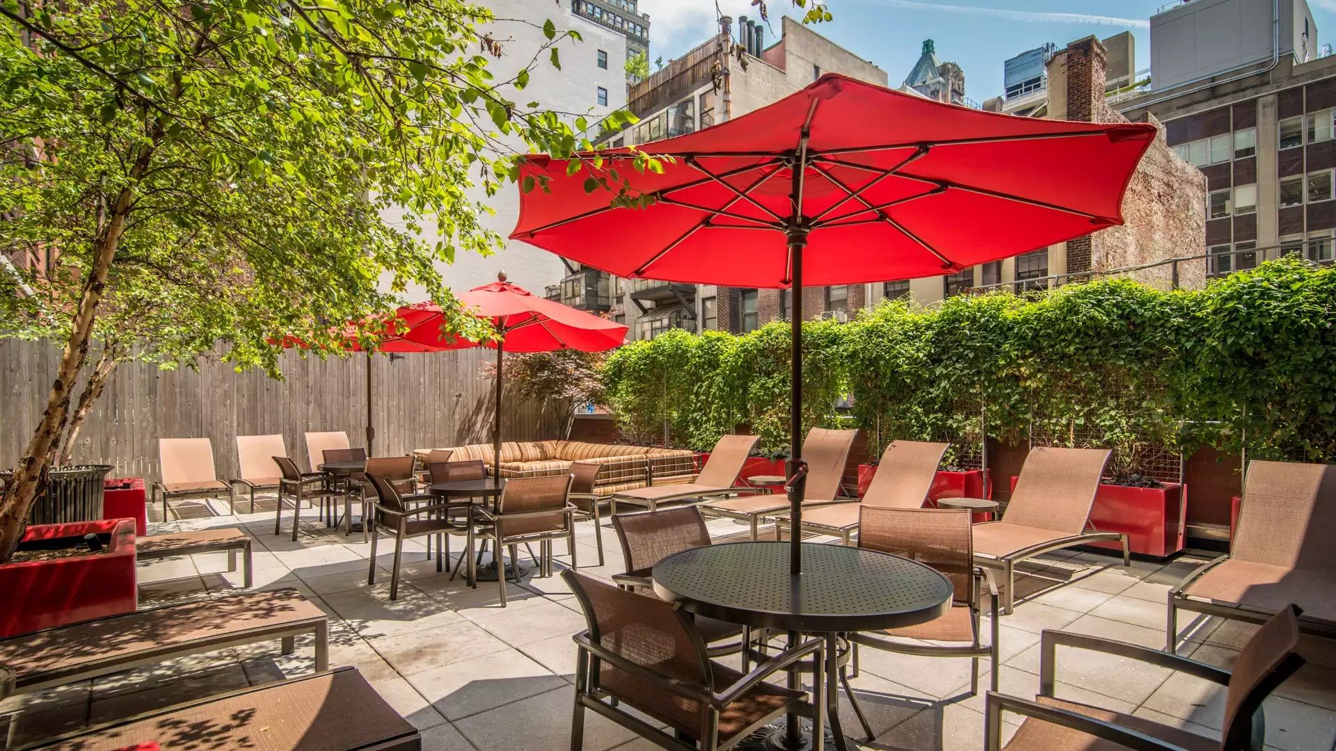 An outdoor patio with tables and chairs, red umbrellas, and a wooden fence.