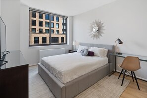 Ein modernes Schlafzimmer mit einem großen Bett, einem Schreibtisch, einem Sessel, einer Stehlampe und Blick auf ein Gebäude durch ein Fenster.