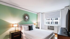 Une chambre bien rangée, avec un lit, des tables de chevet, une télévision et une fenêtre équipée de stores.