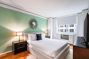 Een keurig opgemaakt slaapkamer met een bed, nachtkastjes, een televisie en een raam met jaloezieën.