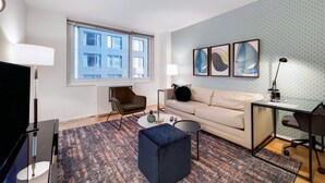 Un salon moderne doté d’une grande fenêtre, d’un canapé, d’une table basse, d’un téléviseur et d’un bureau avec une chaise.
