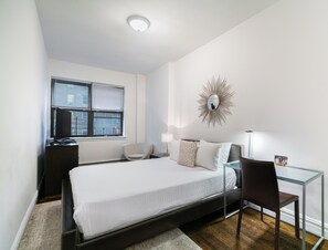 Ein Schlafzimmer mit einem Bett, einem Schreibtisch, einem Stuhl, einem Glastisch, einem Spiegel und einem Fenster mit Jalousien.