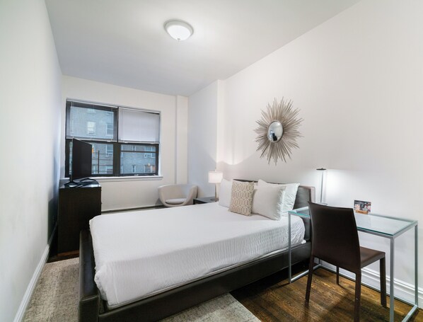 A bedroom with a bed, a desk, a chair, a glass table, a mirror, and a window with blinds.