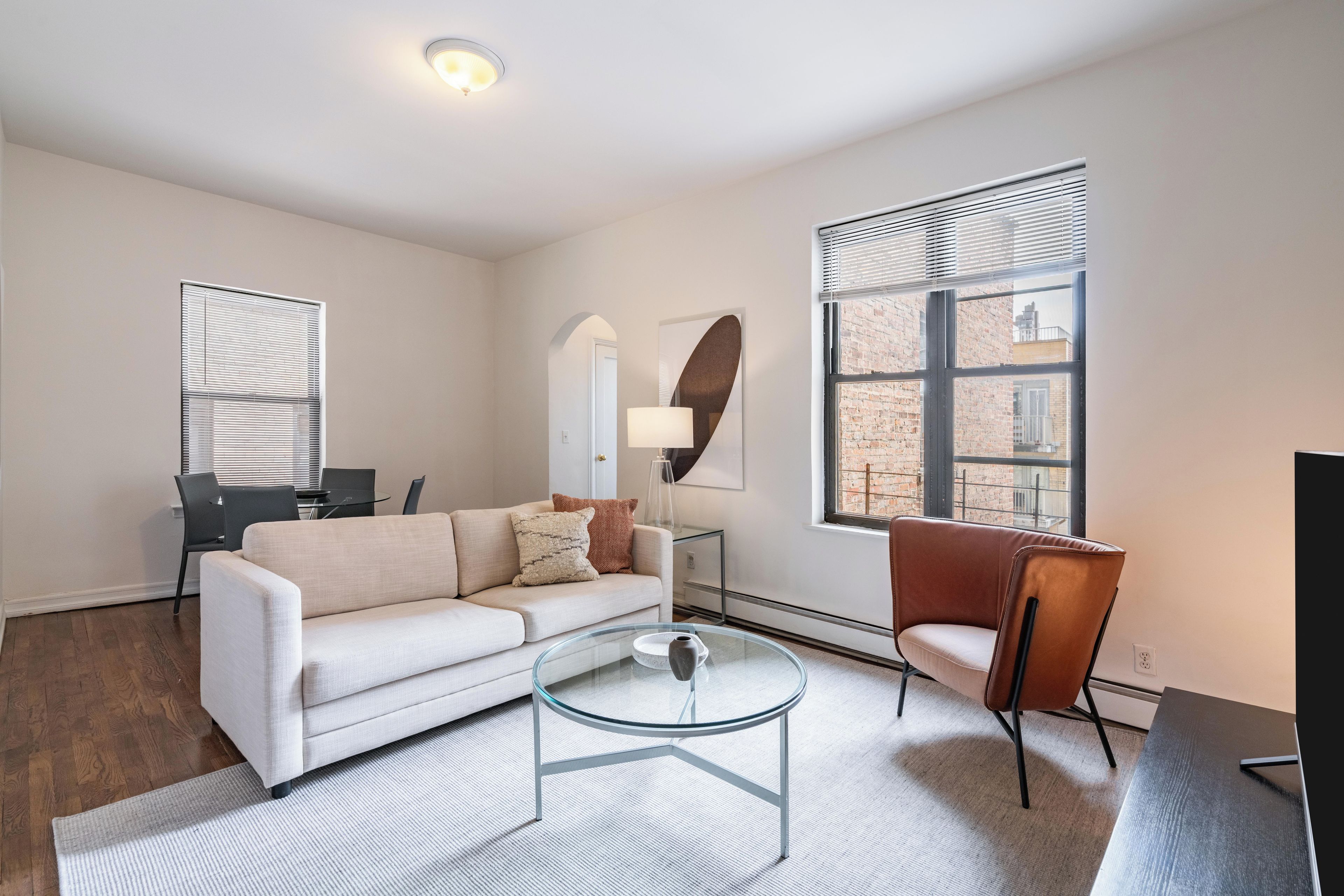 A modern living room with a sofa, a glass coffee table, a chair, a TV, and a window with blinds.