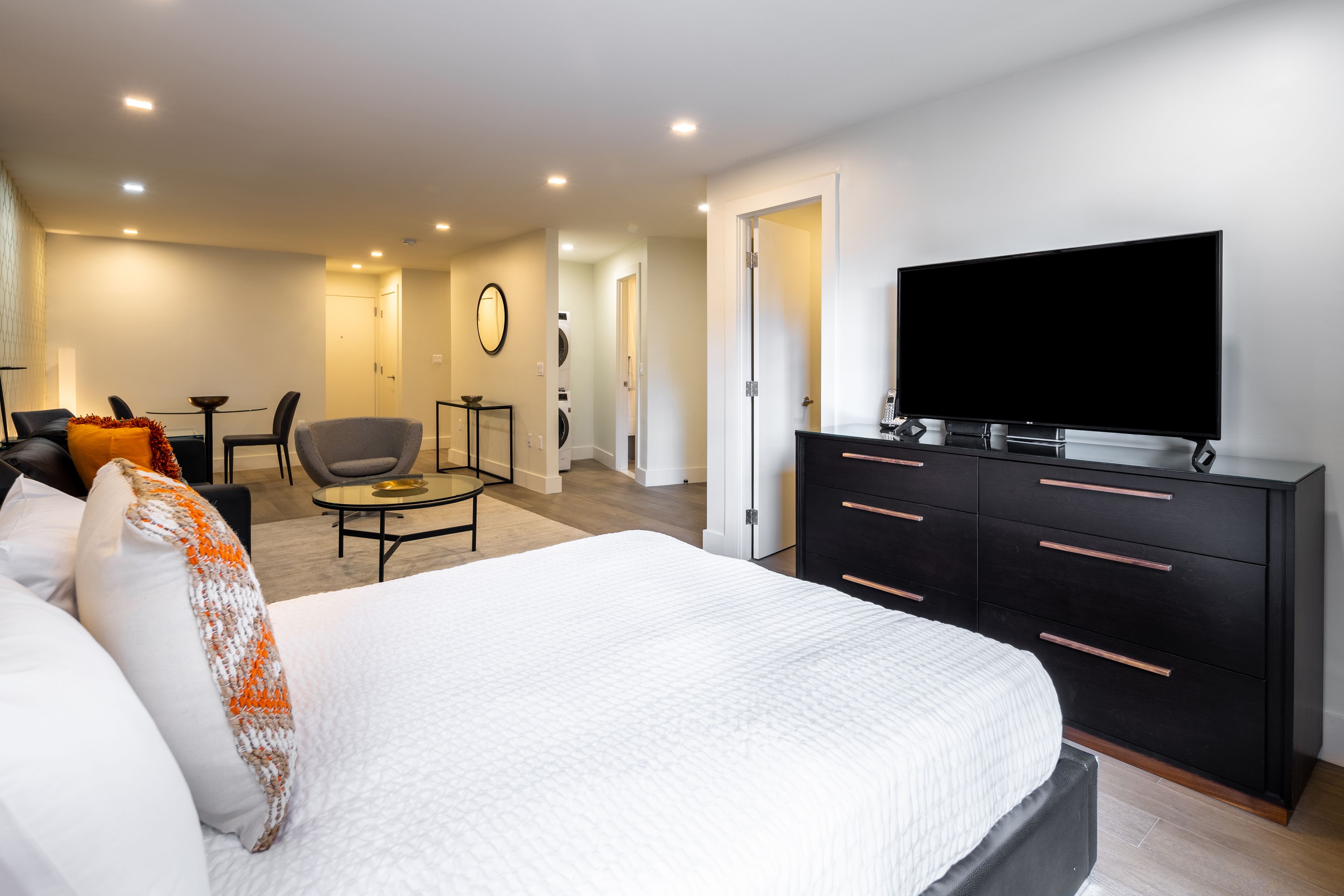 A modern hotel room with a large bed, a flat-screen TV, a black dresser, a round coffee table, and a seating area with a sofa and chairs.