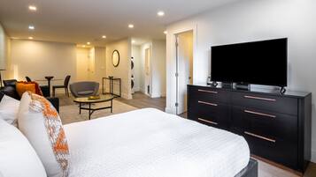 A modern hotel room with a large bed, a flat-screen TV, a black dresser, a round coffee table, and a seating area with a sofa and chairs.
