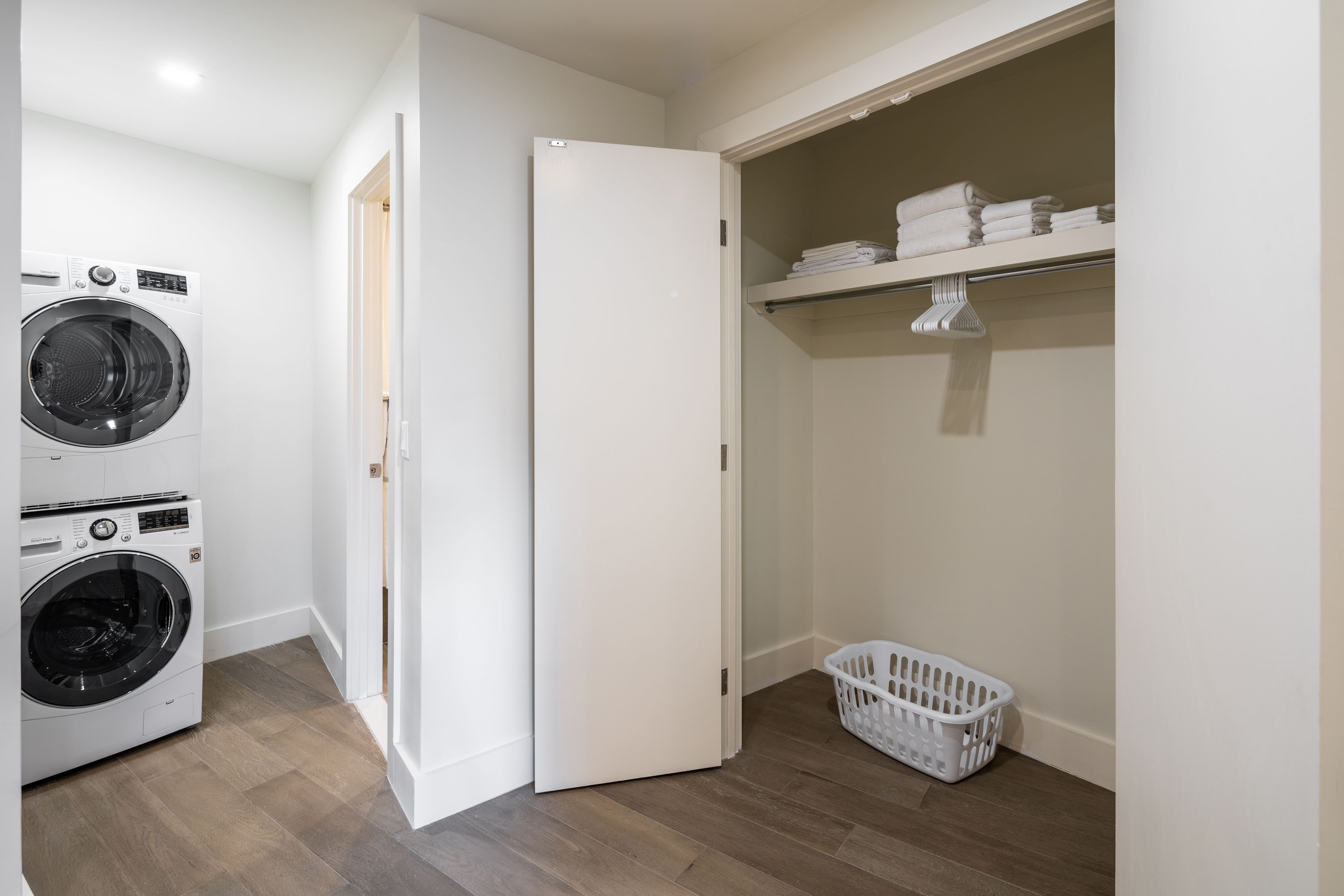 A laundry room with two washing machines, a dryer, a white door, a shelf with folded towels, and a laundry basket.