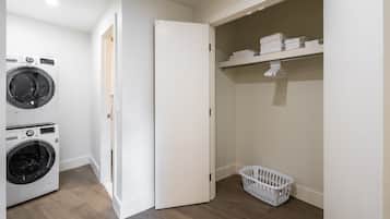 A laundry room with two washing machines, a dryer, a white door, a shelf with folded towels, and a laundry basket.
