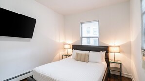 Ein Hotelzimmer mit einem Bett, zwei Nachttischlampen, einem Wand-Fernseher und einem Fenster mit Jalousien.