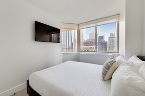 A bedroom with a large bed, a flat-screen TV mounted on the wall, and a window with a city view.