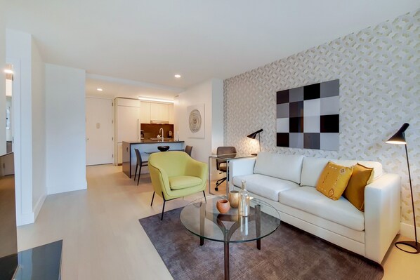 A modern living room with a glass coffee table, a white sofa with yellow cushions, a yellow armchair, and a patterned wall.
