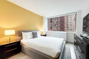 Una habitación de hotel moderna con una cama grande, mesitas de noche con lámparas, un tocador y un ventanal con vistas a la ciudad.
