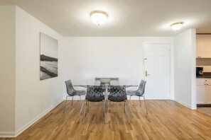 Uma sala de jantar com mesa de vidro e quatro cadeiras, uma porta branca, e área de cozinha visível à direita.