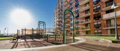 A playground with equipment, a brick pathway, and a multi-story residential building with balconies.