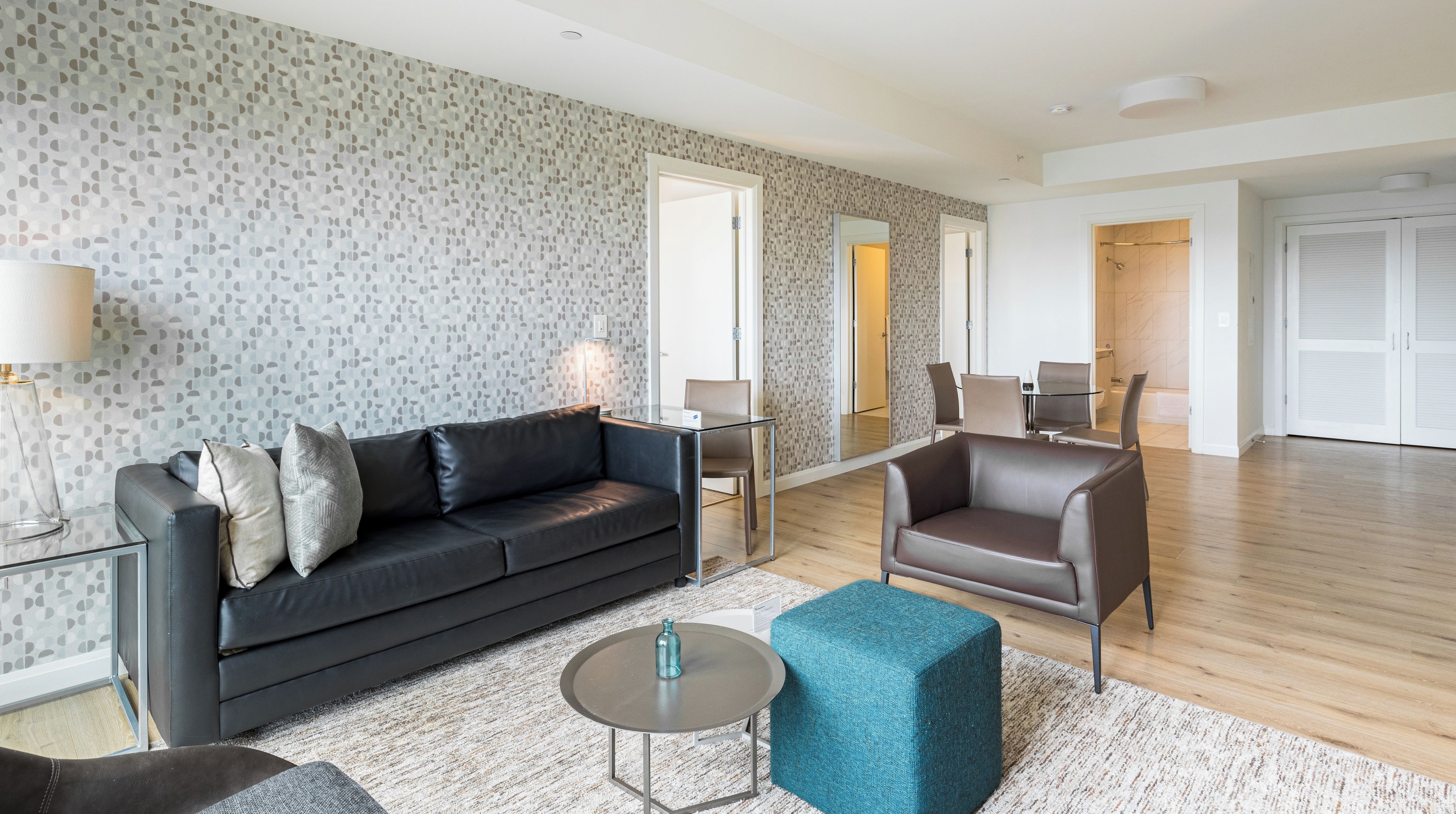 A modern living room with a black leather sofa, a glass coffee table, and a teal ottoman. There is a dining area with a glass table and chairs, and a bathroom with a shower and toilet visible.