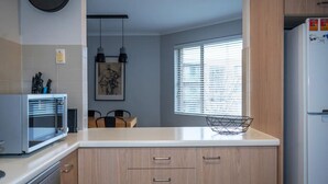 A compact kitchen with a microwave, oven, and refrigerator. There is a window with blinds, a hanging light fixture, and a small wire basket on the counter.