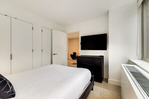 Quarto com cama, guarda-roupa, escrivaninha, cadeira e televisão fixada na parede.