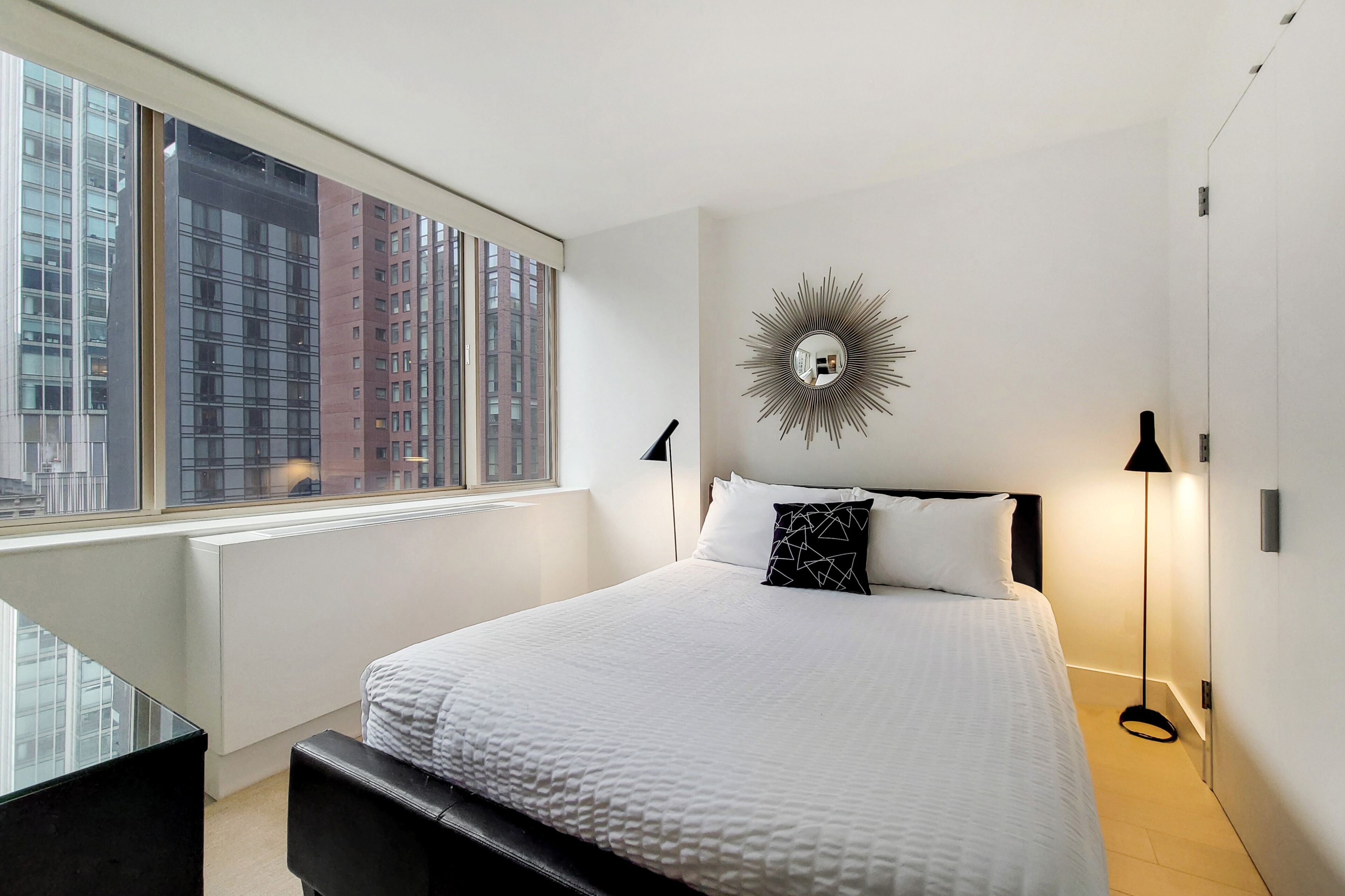 A modern hotel room with a large window, a bed with white bedding, a sunburst mirror on the wall, and a standing floor lamp.