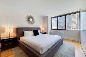 Ein modernes Schlafzimmer mit einem großen Bett, zwei Nachttischen, einem dekorativen Spiegel und Blick auf hohe Gebäude durch ein großes Fenster.
