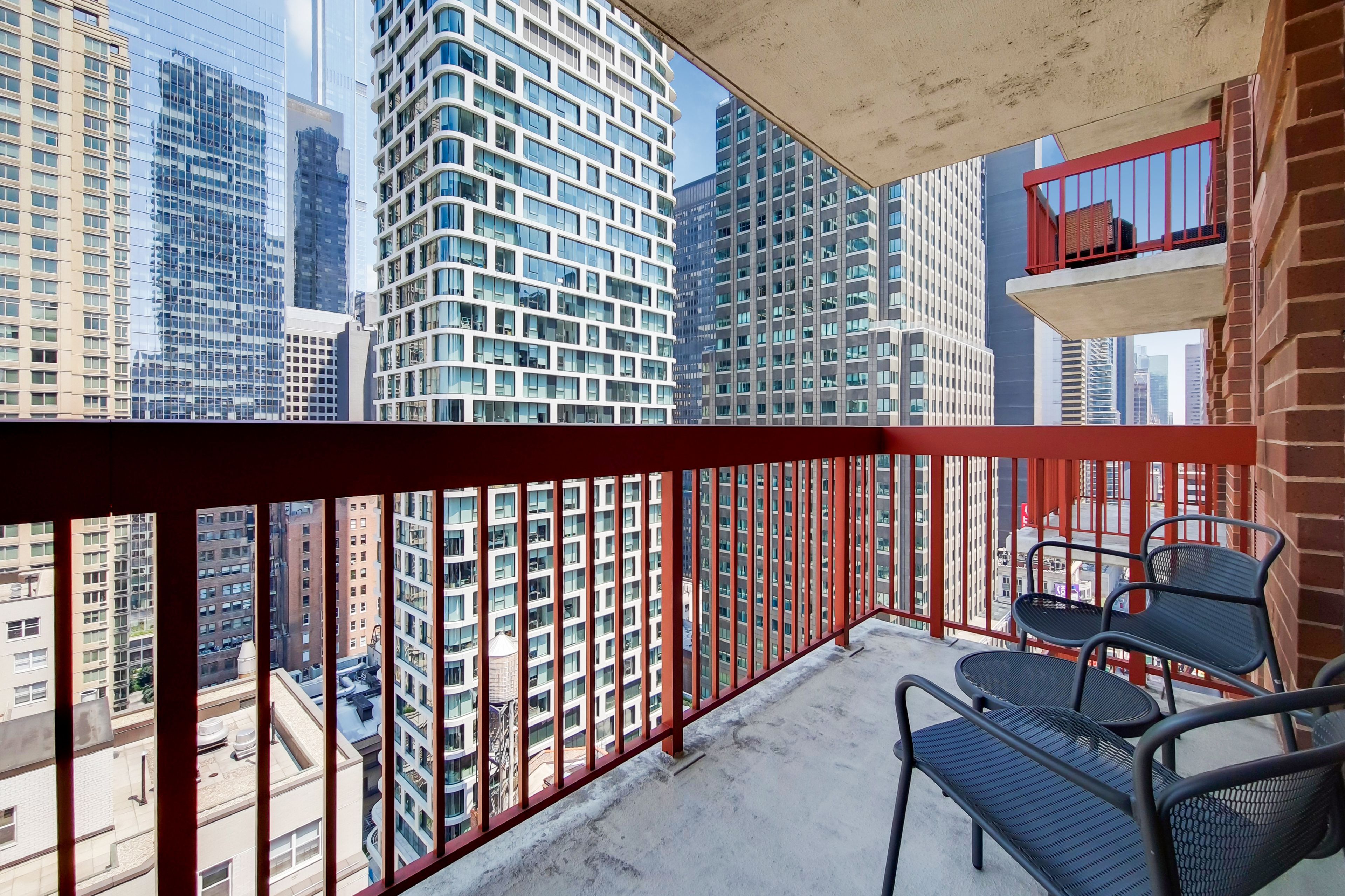 A balcony with red railings, a view of tall buildings, and two black metal chairs.