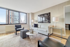 Ein modernes Wohnzimmer mit einem weißen Sofa, einem schwarz-weißen Hocker und einem Glastisch. Ein großes Fenster bietet Blick auf eine Stadtlandschaft.