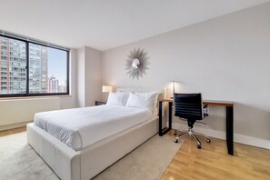 A modern hotel room with a large bed, a desk with a chair, a wall-mounted mirror, and a view of tall buildings.