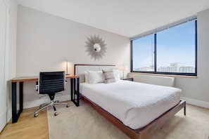Ein modernes Hotelzimmer mit einem großen Bett, einem Schreibtisch, einem Sessel, einem Nachttisch, einer Lampe und Blick auf die Stadt durch ein großes Fenster.