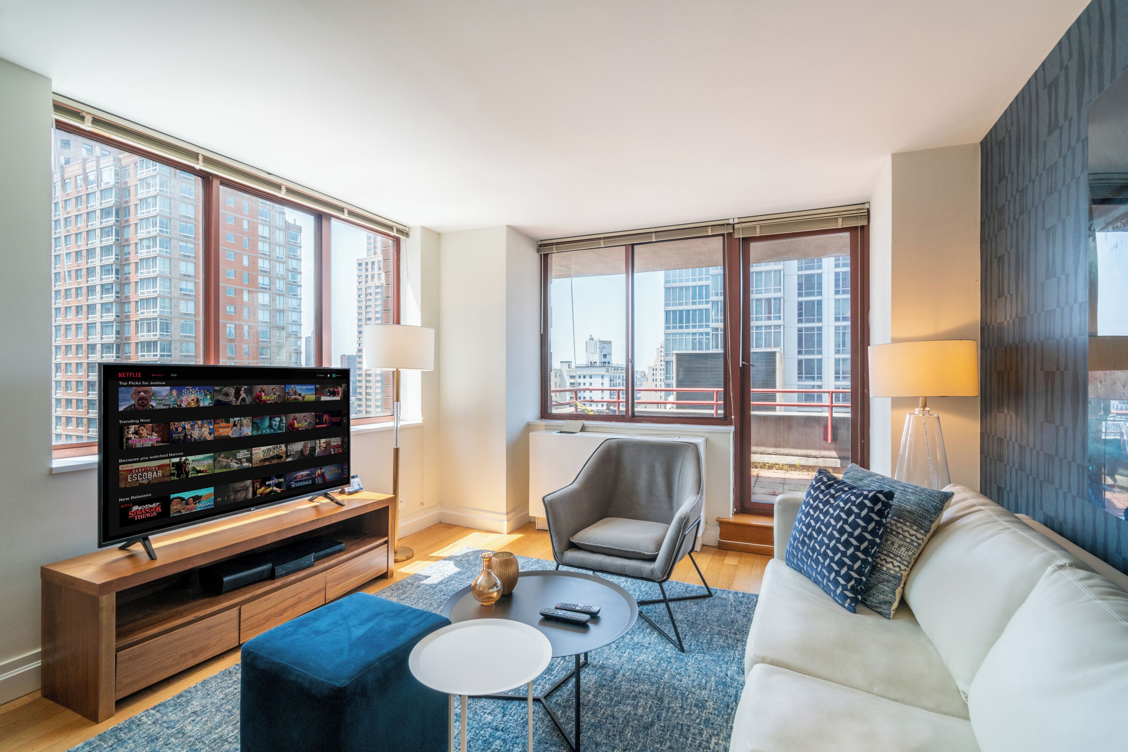 A modern living room with a flat-screen TV, comfortable seating, and large windows offering city views.