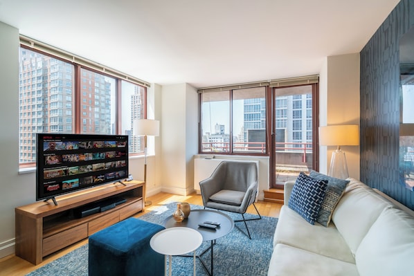 A modern living room with a flat-screen TV, comfortable seating, and large windows offering city views.