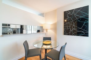 A modern kitchen with white cabinets and a built-in microwave, a glass dining table with two chairs, and a contemporary abstract painting on the wall. - Gorgeous One Bed in Midtown West Manhattan (New York)