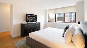 A modern bedroom with a large bed, a TV on a dresser, a window with a city view, and a door leading to another room. - Gorgeous One Bed in Midtown West Manhattan (New York)