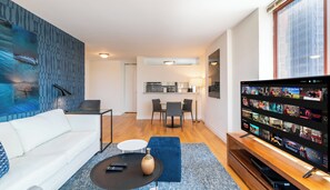 A modern living room with a white sofa, a coffee table, and a TV displaying a Netflix interface. A kitchen is visible in the background. - Luxury Midtown Living | Balcony & Fitness Center (New York)