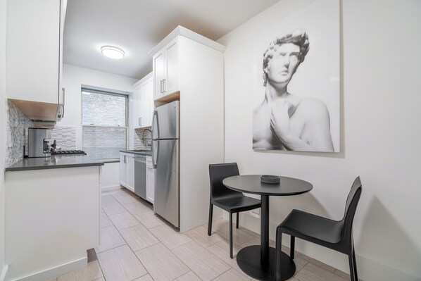A modern kitchen with white cabinets, a stainless steel refrigerator, a window, and a dining area with a table and chairs.