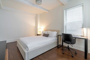 A bedroom with a bed, a desk, a chair, a lamp, and a window with blinds.